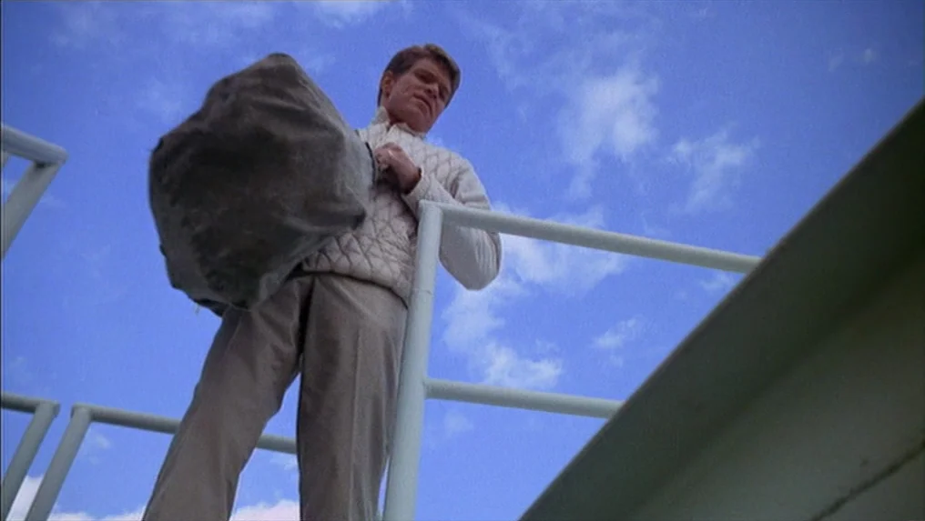 Targets (1968) movie still: fullbody — A man descends a simple white metal staircase set against a clear blue sky. He holds a large, grey, shapeless…