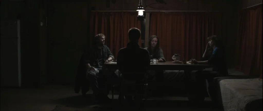 One Hundred Mornings (2009) movie still: wide — A dimly lit room with wood-paneled walls and red curtains behind a table. Four people sit around the…