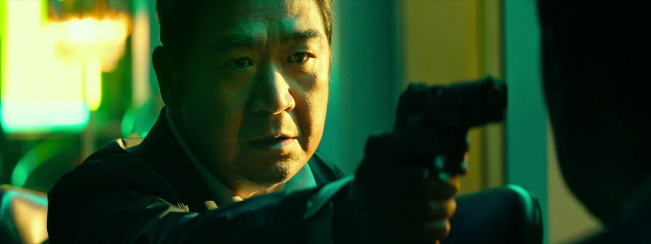 Under The Light (2023) movie still: closeup — An office interior at night bathed in strong green light. A man holds a pistol directed toward a…
