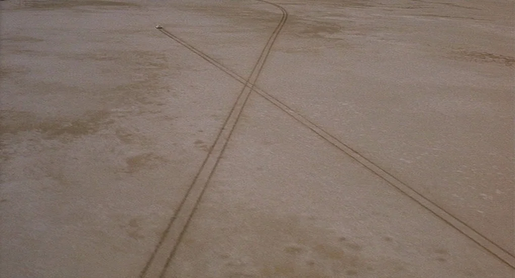 Vanishing Point (1971) movie still: establishing — A vast, flat, and empty desert landscape shows pale, parched earth. A white vehicle travels in…