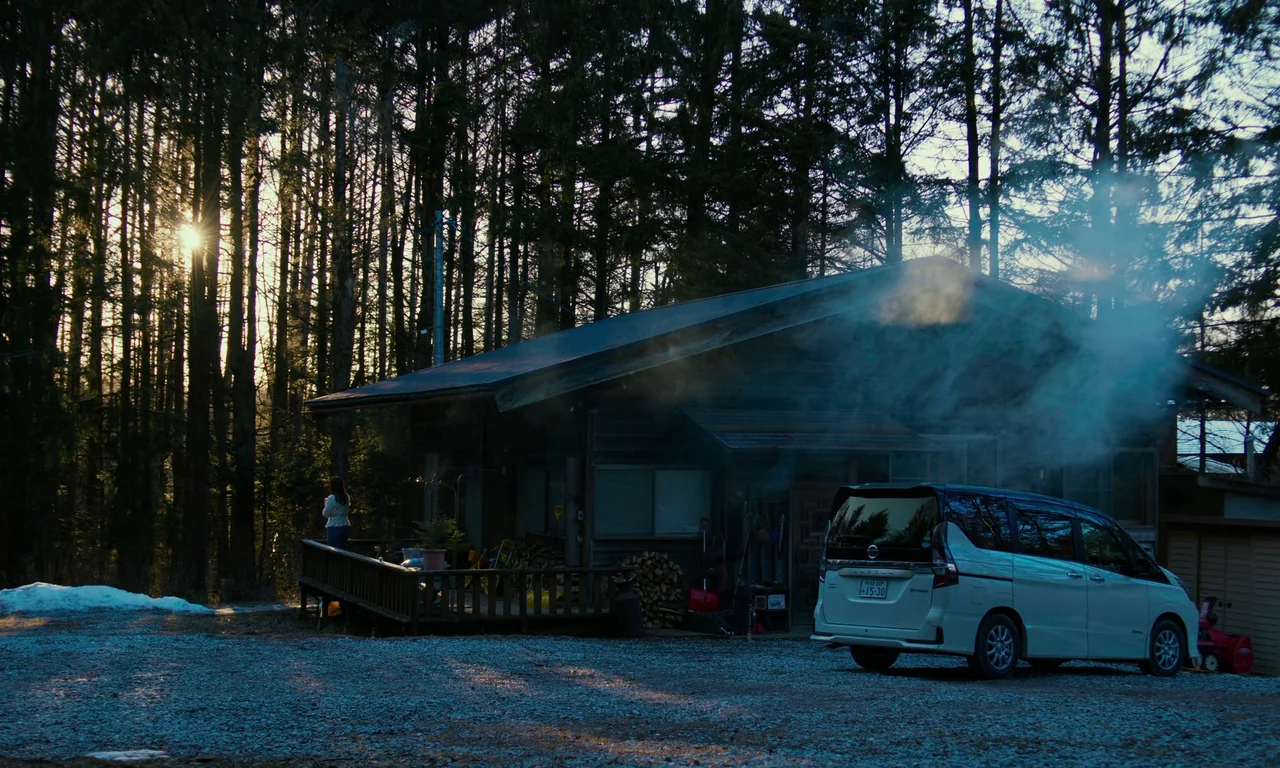 Evil Does Not Exist (2023) movie still: wide — A wooden cabin sits at the edge of a dense forest during late afternoon. A white minivan is parked on…