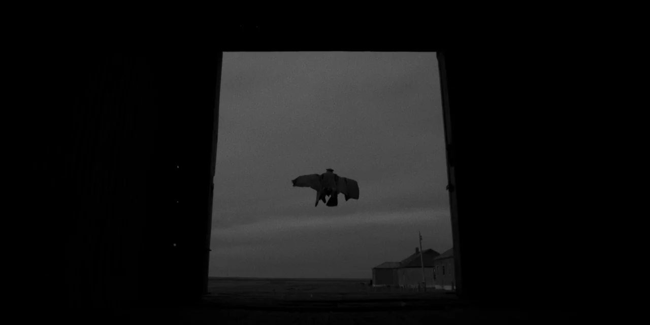 El Conde (2023) movie still: wide — An open landscape under an overcast sky viewed from a dark building interior. A man wearing a coat jumps in the…