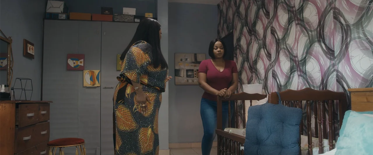 Big Love (2012) movie still: medium — A bedroom features patterned wallpaper, a wooden crib, and a dresser against the wall. An older woman in a…