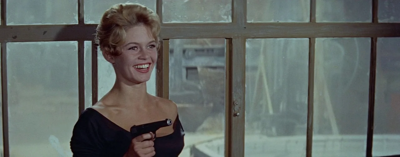 And God Created Woman (1956) movie still: medium — A woman stands before a large, multi-paned window inside an industrial workshop structure. She…
