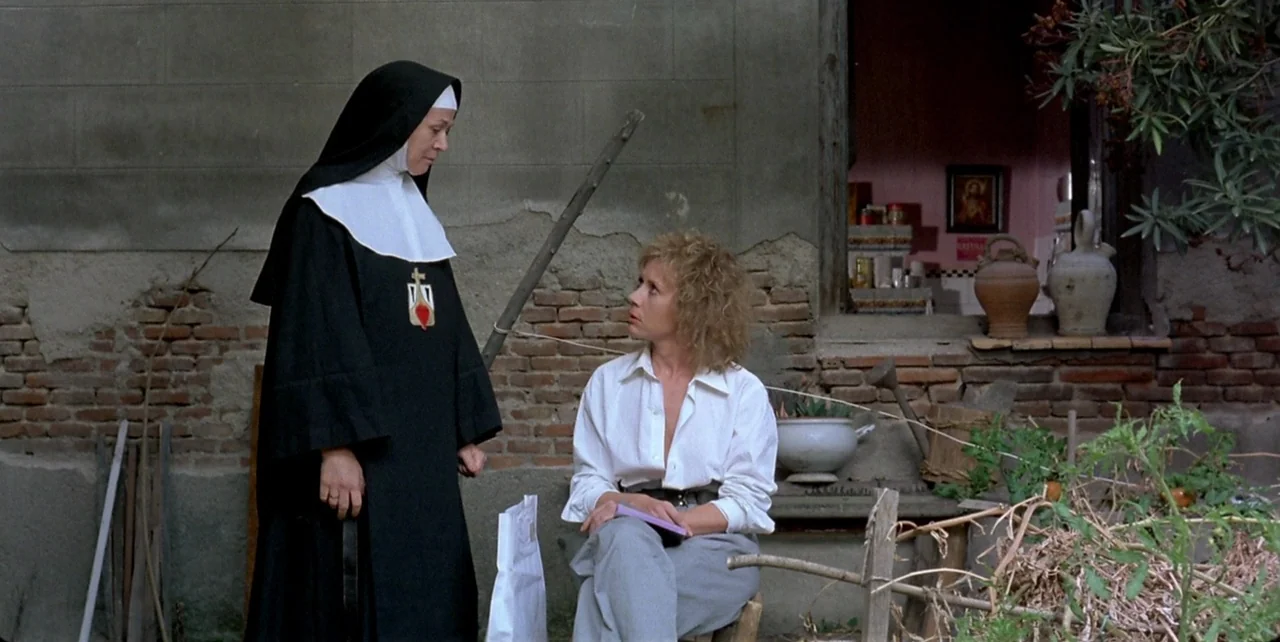 Dark Habits (1983) movie still: fullbody — A rural courtyard features a weathered brick wall and a small windowed opening. A nun faces a blonde…