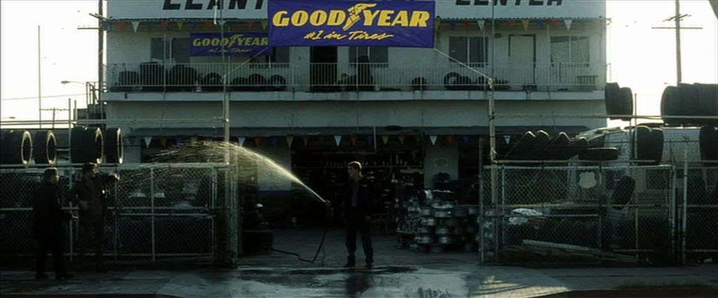 Fight Club (1999) movie still: wide — A two-story commercial building with stacked tires and blue Goodyear banners on the facade. Three men stand in…