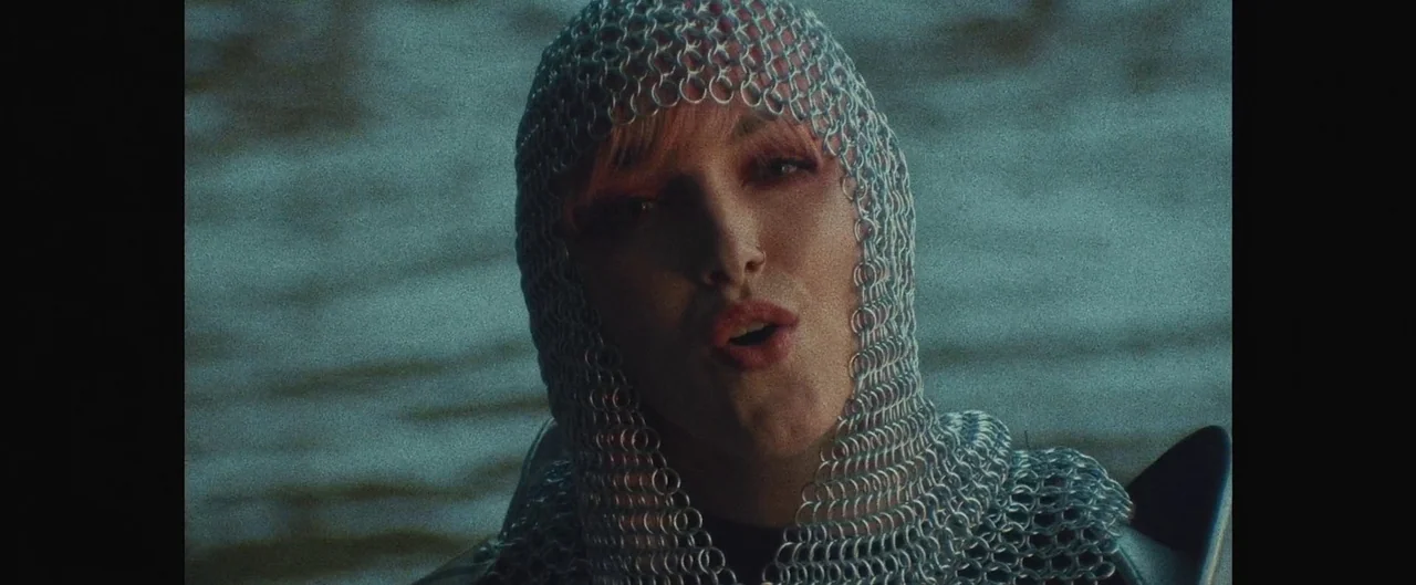 Poser (2021) movie still: closeup — A dark, blurred water surface creates a textured backdrop behind the subject. A person wears a metallic…