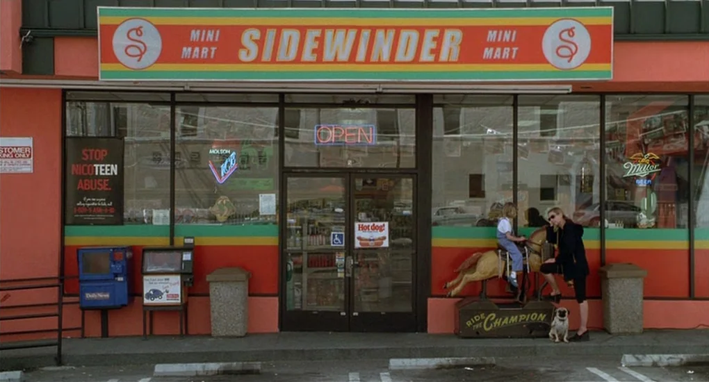 Ghost World (2001) movie still: wide — A 1990s convenience store exterior features red walls and a large, colorful neon-trimmed sign. A small child…