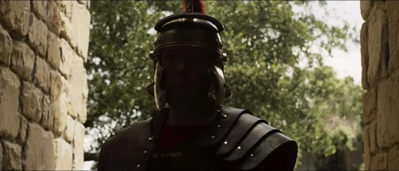 The Penitent Thief (2021) movie still: medium — A Roman soldier stands within a textured stone archway under natural daylight conditions. The…