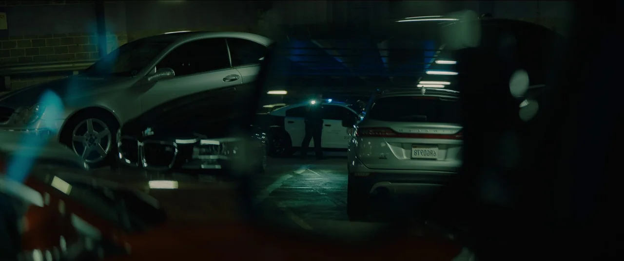 Night Teeth (2021) movie still: wide — A dimly lit concrete parking garage features several parked cars and overhead fluorescent lighting rows. An…