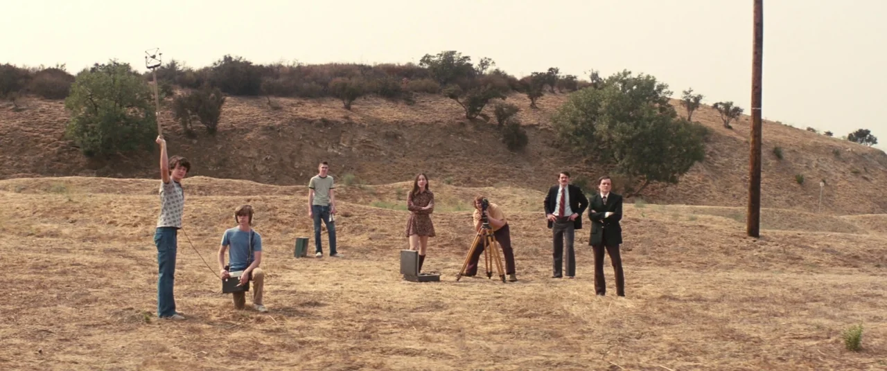 Licorice Pizza (2021) movie still: wide — A dry, grassy hillside with sparse shrubs beneath a pale, overcast sky. A group of young people and two…