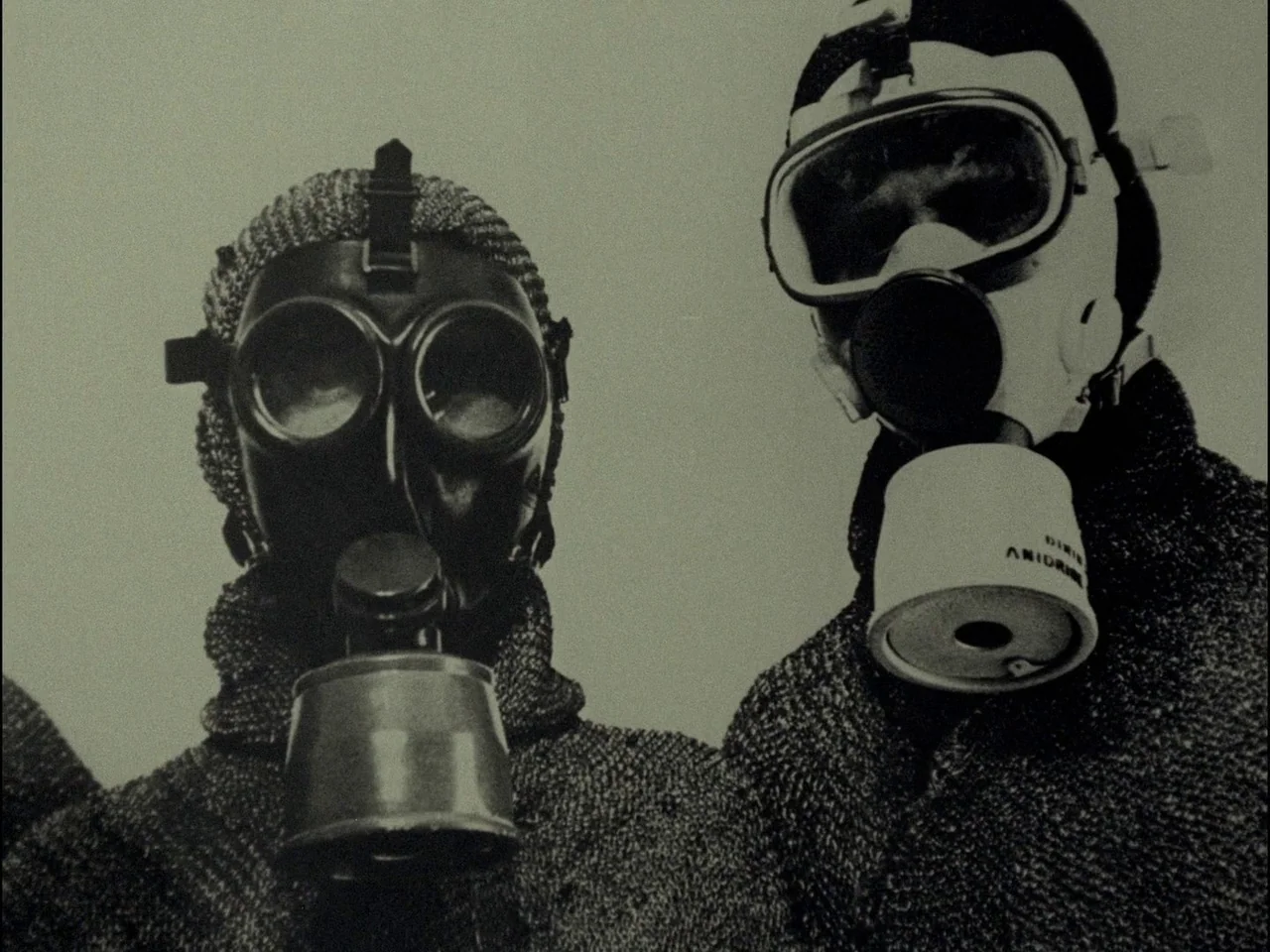 Dillinger is Dead (1969) movie still: medium — Two individuals stand against a featureless sky wearing varied industrial-style respirators and rough…