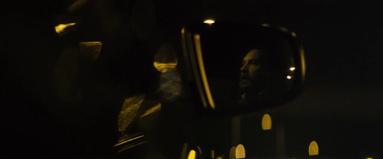 Locke (2013) movie still: closeup — A side view car mirror at night reflects a man sitting in the driver's seat. Golden out-of-focus lights blur…
