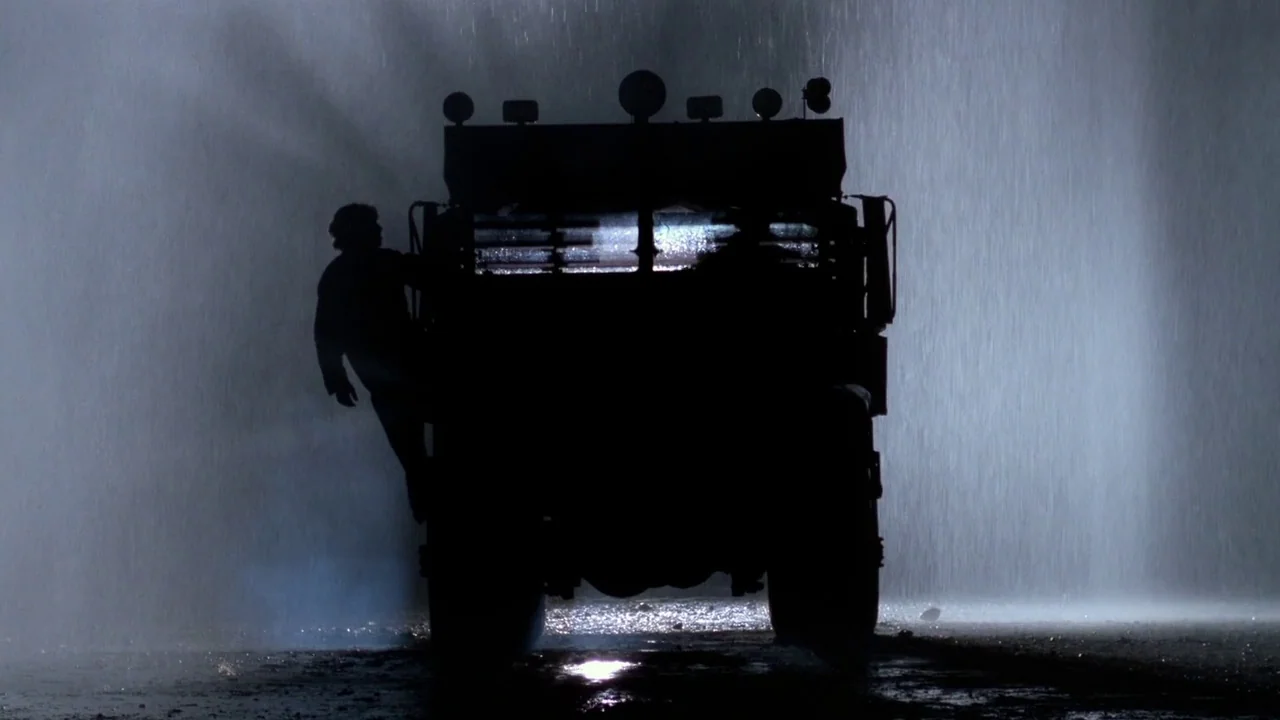 Sorcerer (1977) movie still: wide — A heavy industrial truck drives down a dark road during a torrential rainstorm. A man hangs onto the side of the…