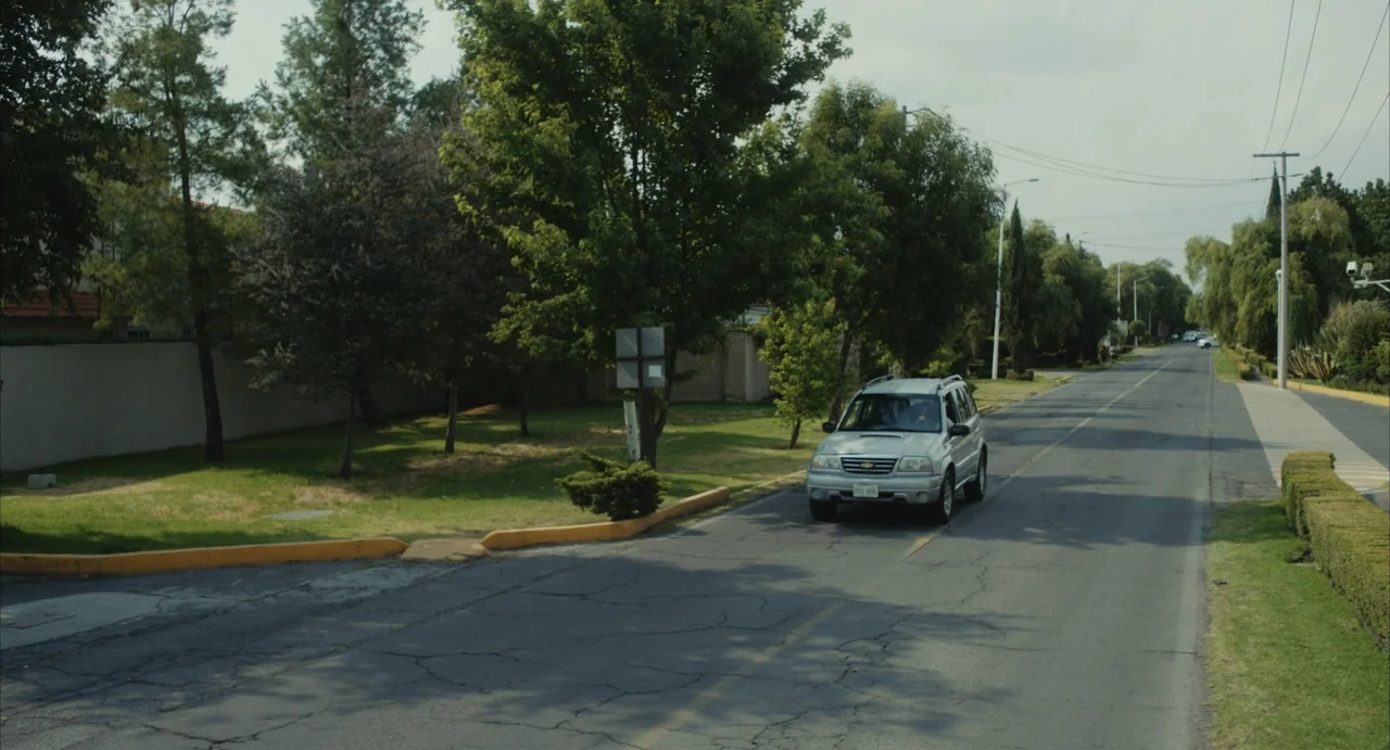 The Diary (1999) movie still: wide — A suburban street lined with green trees and manicured lawns under a bright sky. A silver SUV travels along the…