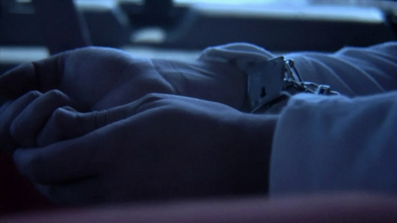 A Horrible Way To Die (2010) movie still: closeup — A close view of hands cuffed together inside a dimly lit vehicle. Metal restraints bind the…
