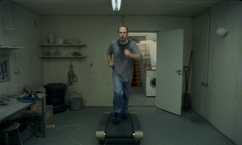 You, The Living (2007) movie still: fullbody — A utilitarian garage storage space with concrete floors and shelves holding assorted mechanical…
