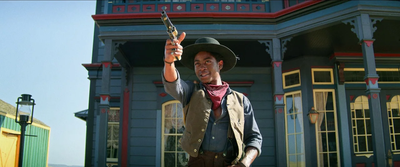 The Harder They Fall (2021) movie still: medium — A man stands before a colorful wooden structure with intricate decorative trim and windows. He…