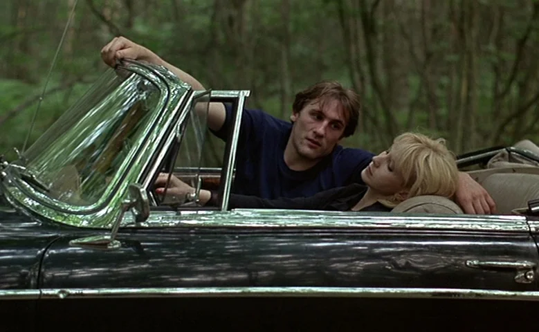 Maîtresse (1975) movie still: medium — A dark vintage convertible sits on a road surrounded by a dense forest. A man with brown hair and a woman…