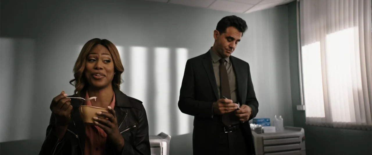 Jolt (2021) movie still: medium — An examination room with pale green walls and vertical window blinds. A woman eats from a bowl while a man in a…