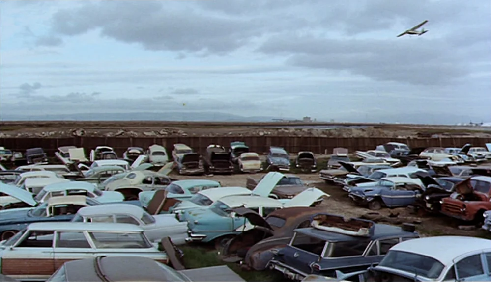 Harold and Maude (1971) movie still: wide — A vast collection of vintage automobiles is parked in a sprawling open-air junkyard area. An airplane…