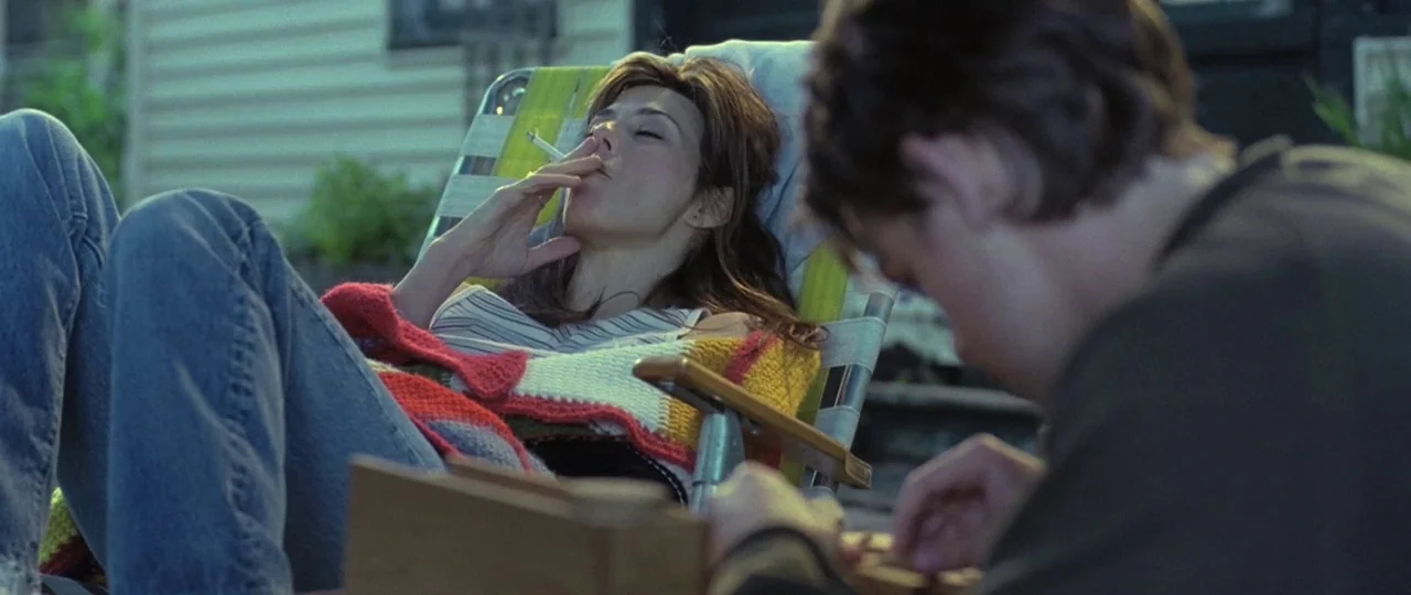 In the Bedroom (2001) movie still: medium — A woman reclines in a folding lawn chair on a residential patio surface. The woman holds a lit cigarette…