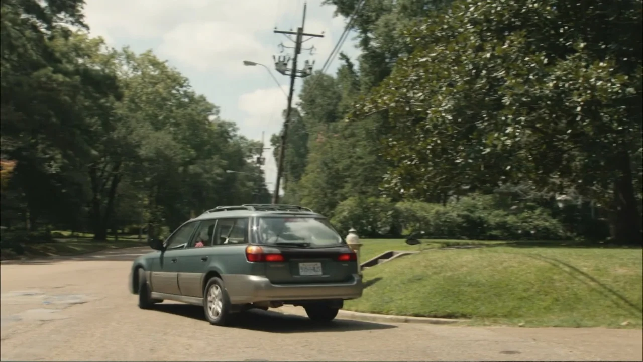 The Hollars (2016) movie still: wide — A suburban street lined with mature green trees on a bright afternoon. A station wagon drives away along the…