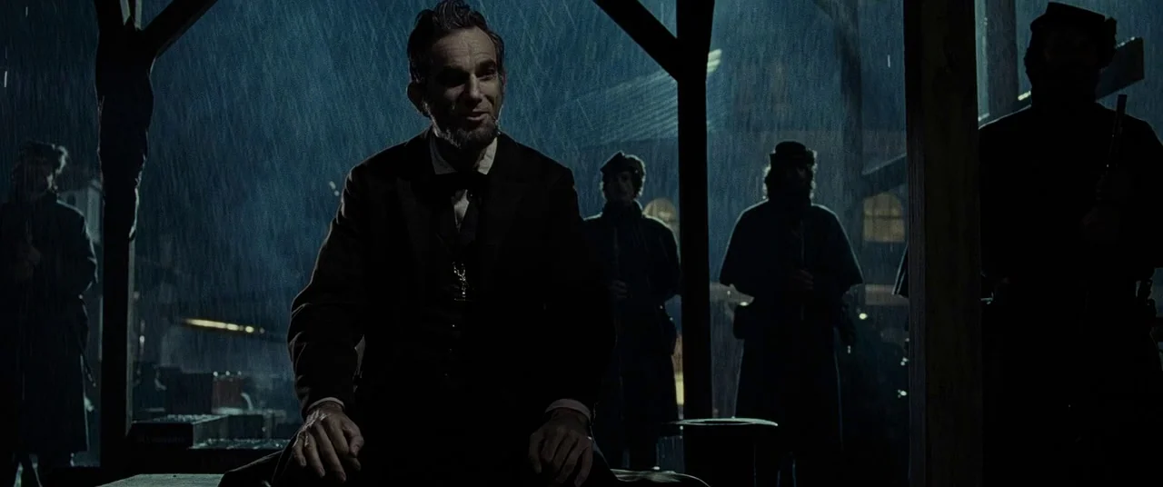 Lincoln (2012) movie still: medium — A dark wooden covered walkway is pelted by heavy rain in the middle of night. Abraham Lincoln sits in the…