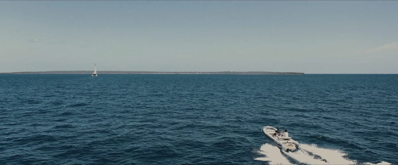 Road House (2024) movie still: wide — A vast expanse of blue ocean extends under a clear, bright daylight sky. A small motorboat moves through the…