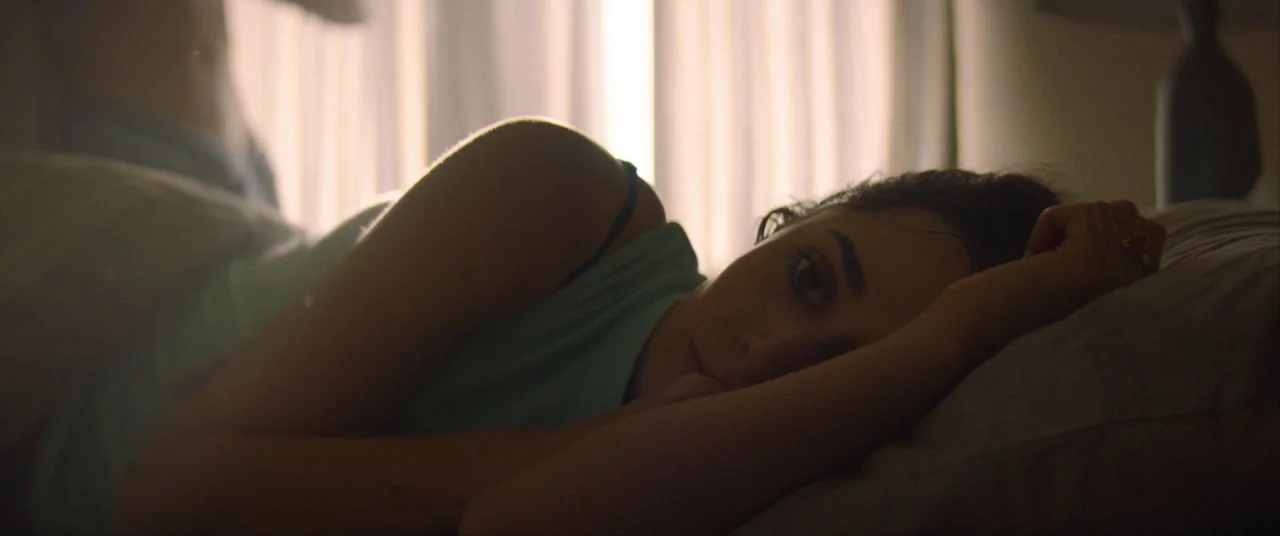 Palm Springs (2020) movie still: closeup — A person lies on a bed with white bedding in front of sheer curtains. The subject rests their head on a…