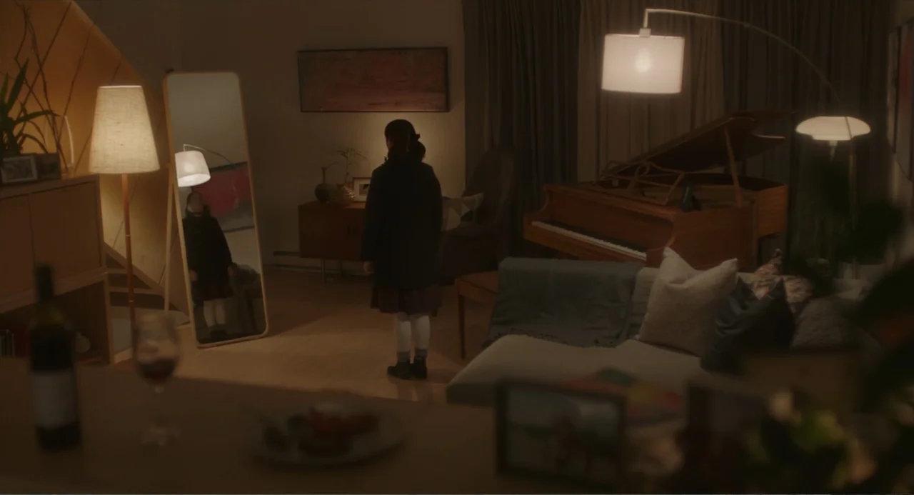 Orphan: First Kill (2022) movie still: medium — A dimly lit room contains a piano, a gray sofa, and a tall mirror. A young girl in a dark coat…