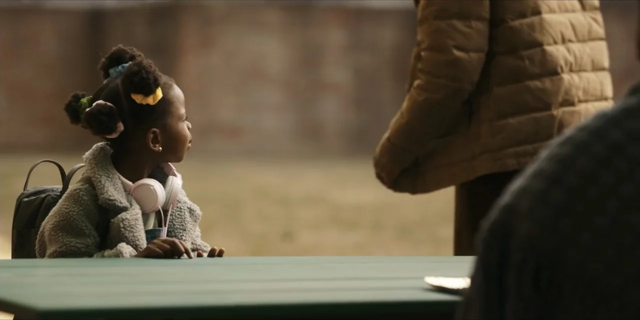 Autumn Beat (2022) movie still: medium — An outdoor park setting with a green table in the foreground. A young girl sits at the table looking at an…