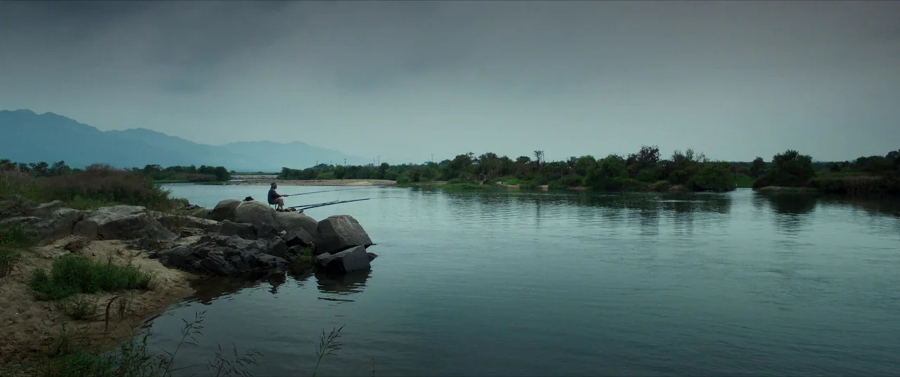 The Wailing (2016) movie still: wide — A wide river winds through a landscape under a heavy, overcast gray sky. A lone person sits on large rocks at…