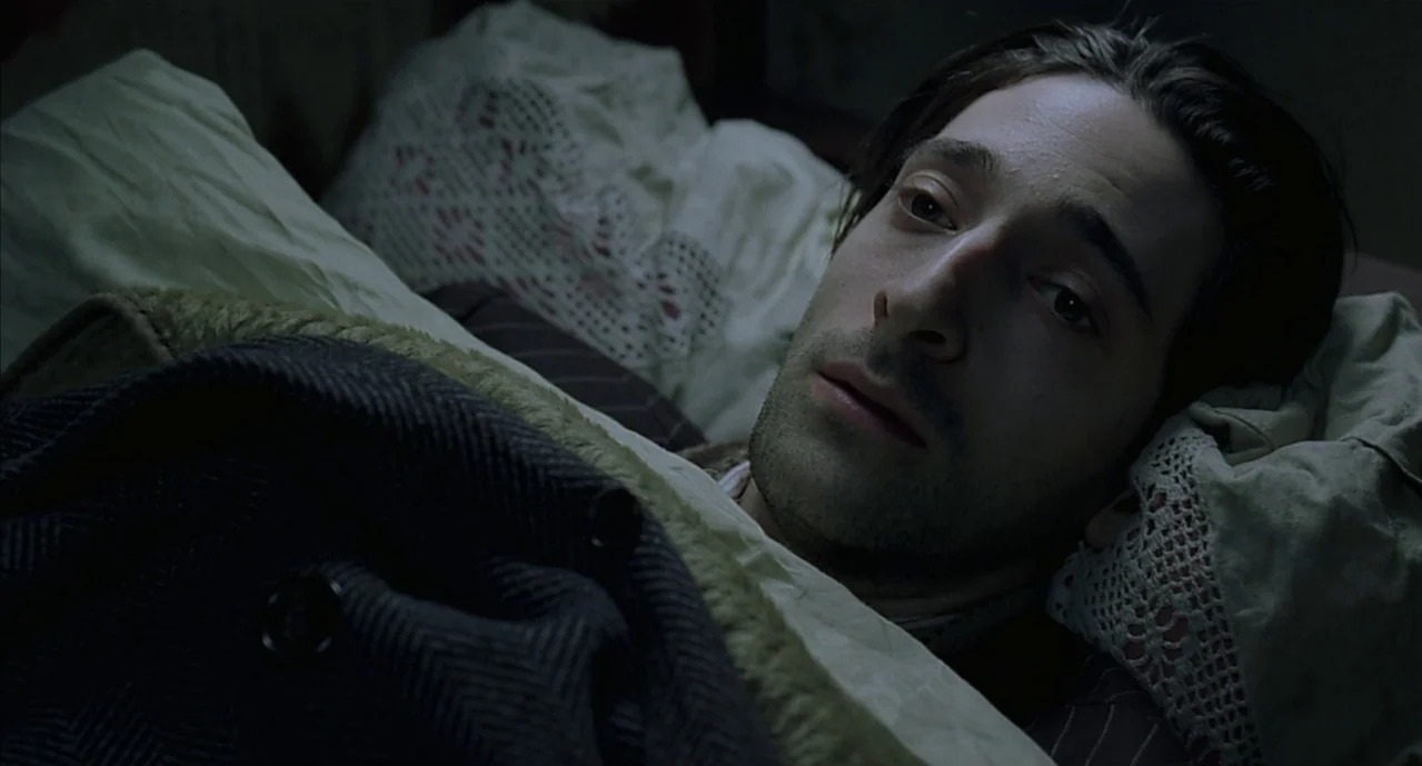 The Pianist (2002) movie still: closeup — A dimly lit room with off-white linens and a crochet pillowcase. A man in a herringbone coat lies resting…