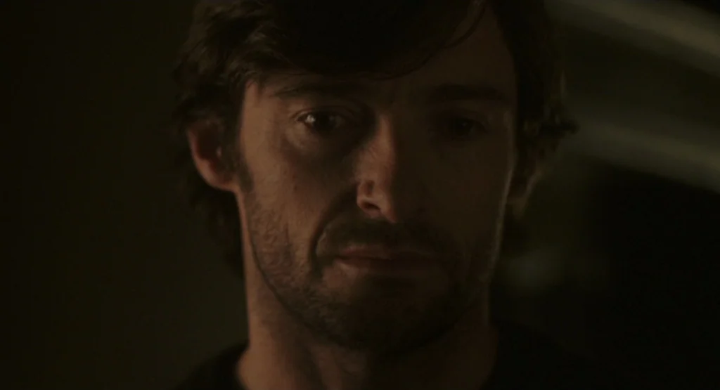 The Fountain (2006) movie still: closeup — A man stands in a dark, dimly lit room with minimal background detail visible. The man looks down with…