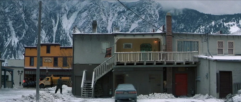 The Pledge (2001) movie still: wide — A quiet town street sits below steep, snow-covered mountains on an overcast winter day. Two-story buildings…