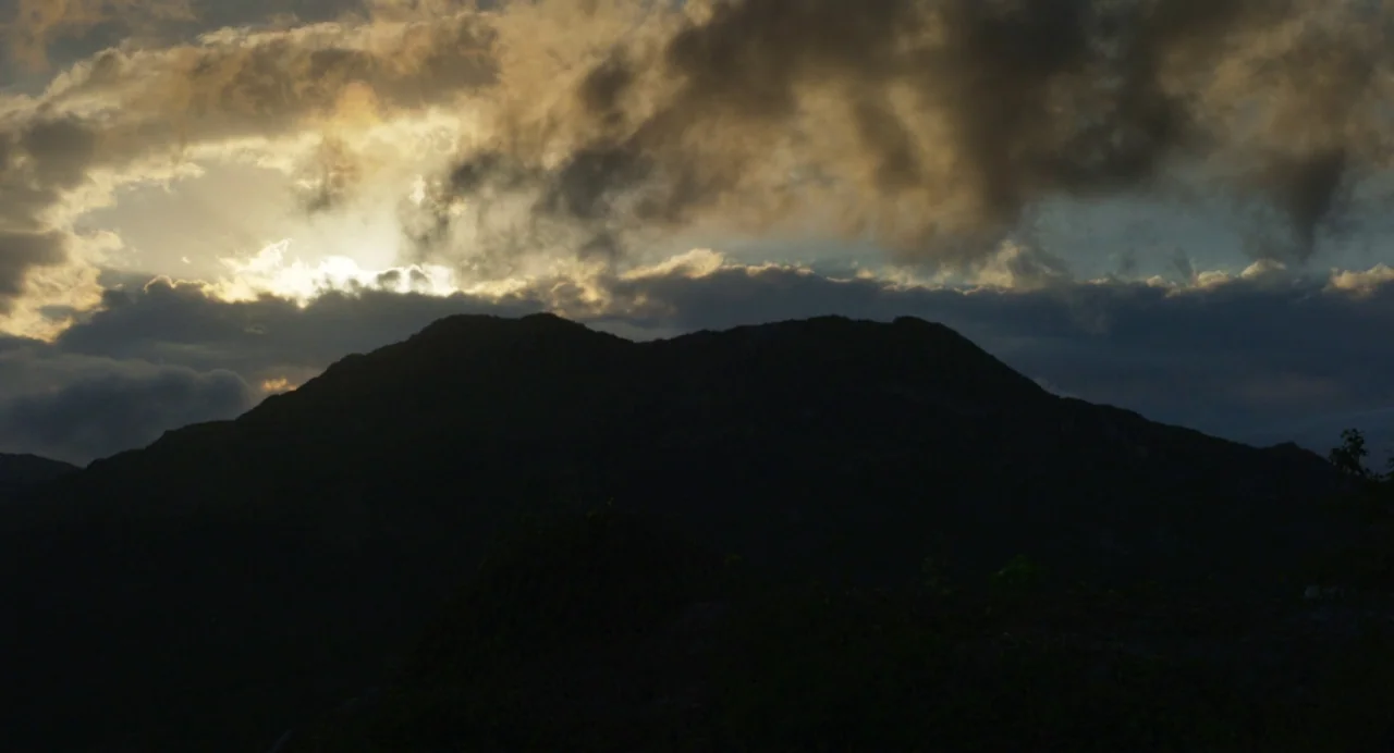 Zombi Child (2019) movie still: establishing — A dark mountain silhouetted against a cloudy sky at sunset. Sun rays penetrate the cloud cover behind…