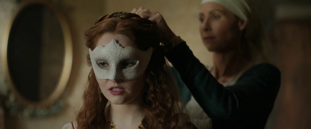 Rosaline (2022) movie still: medium — An interior space contains a woman with red hair wearing a white masquerade mask. A second woman stands behind…