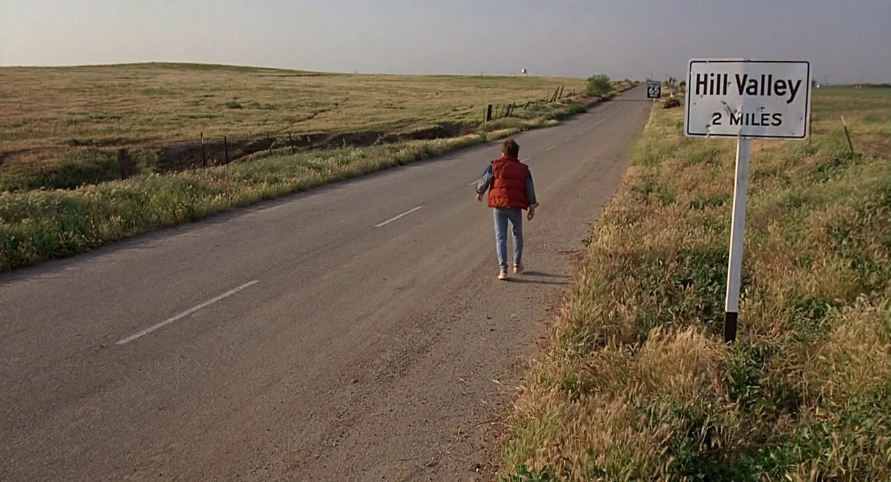Back to the Future (1985) movie still: wide — A desolate rural asphalt road stretches into a grassy field under a pale sky. A lone young man walks…