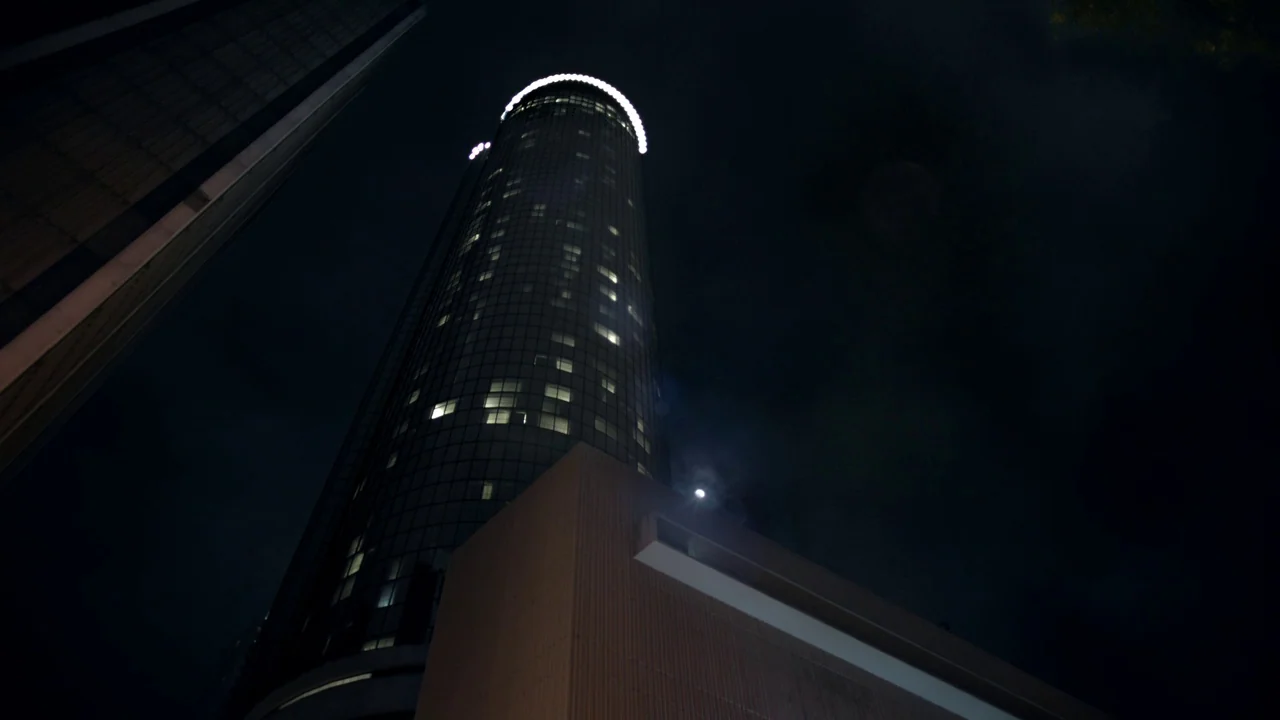 Cult of Chucky (2017) movie still: establishing — A dark, cylindrical skyscraper rises into the night sky with illuminated office windows. The…