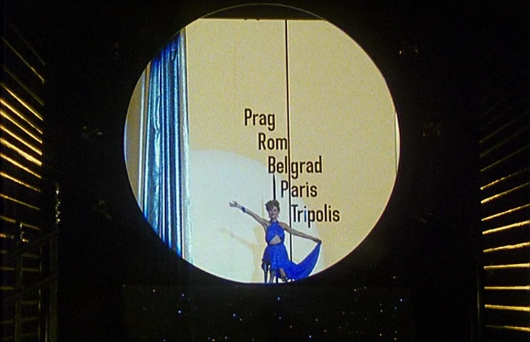 Lili Marleen (1981) movie still: wide — A woman performs on a stage framed within a large circular projection surface. Text lists city names…
