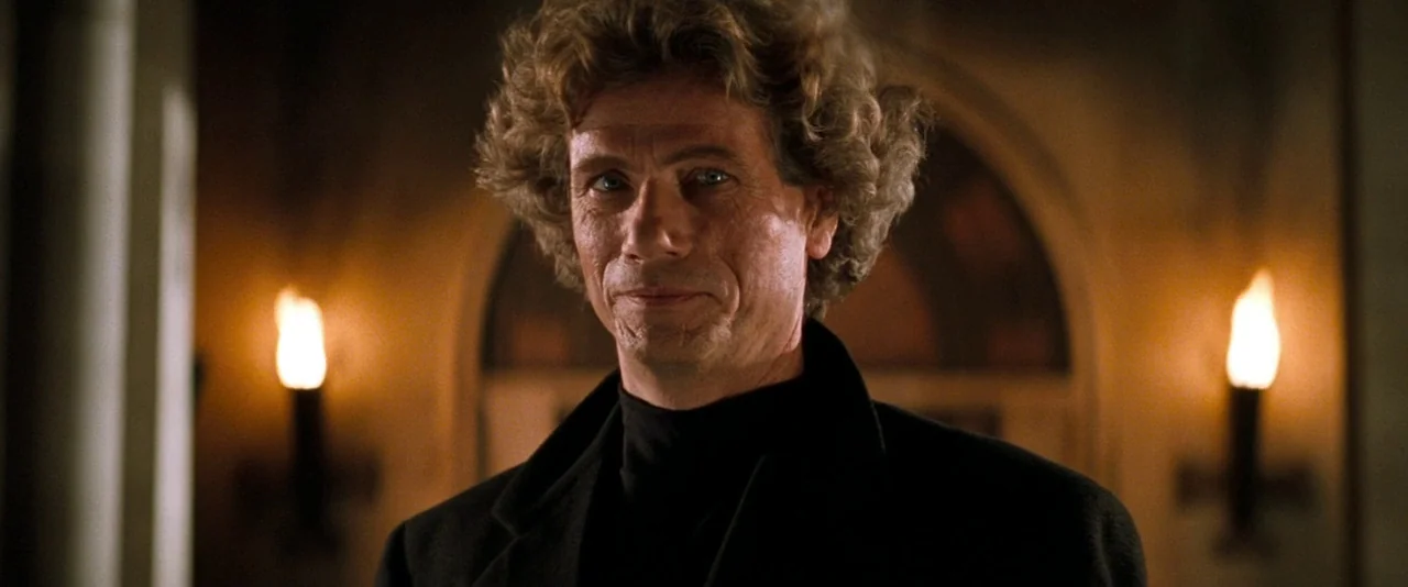In The Mouth of Madness (1994) movie still: closeup — A dimly lit stone corridor features arched alcoves with flickering wall torches. A middle-aged…