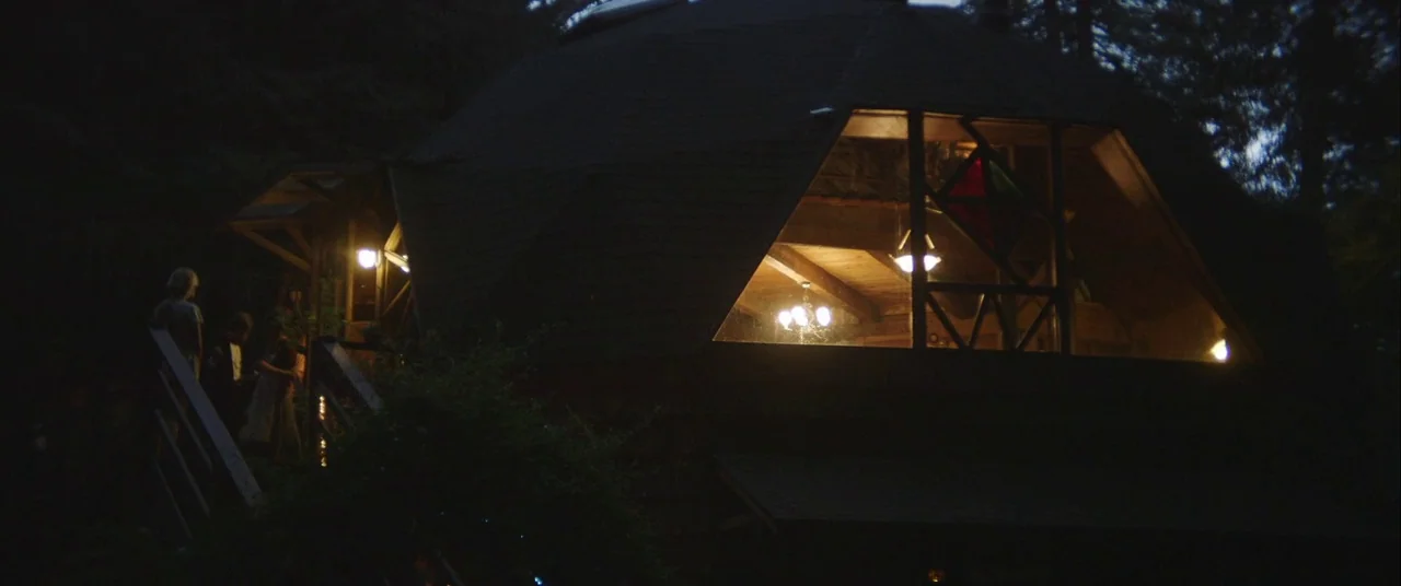 Always Shine (2016) movie still: wide — A dark wooden geodesic dome structure is nestled among trees at night. Warm yellow light shines through the…