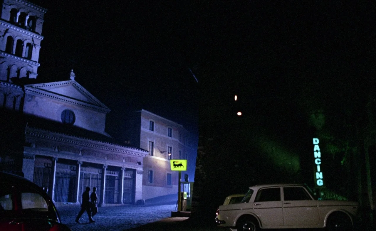 Blood And Black Lace (1964) movie still: wide — An Italian-style piazza at night features a large church with a tall campanile tower. A white sedan…