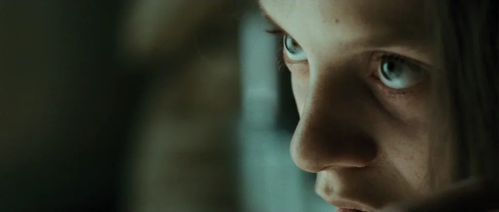 Adam Resurrected (2008) movie still: closeup — A dimly lit interior space with an indistinct, dark background. A young woman's face is partially lit…