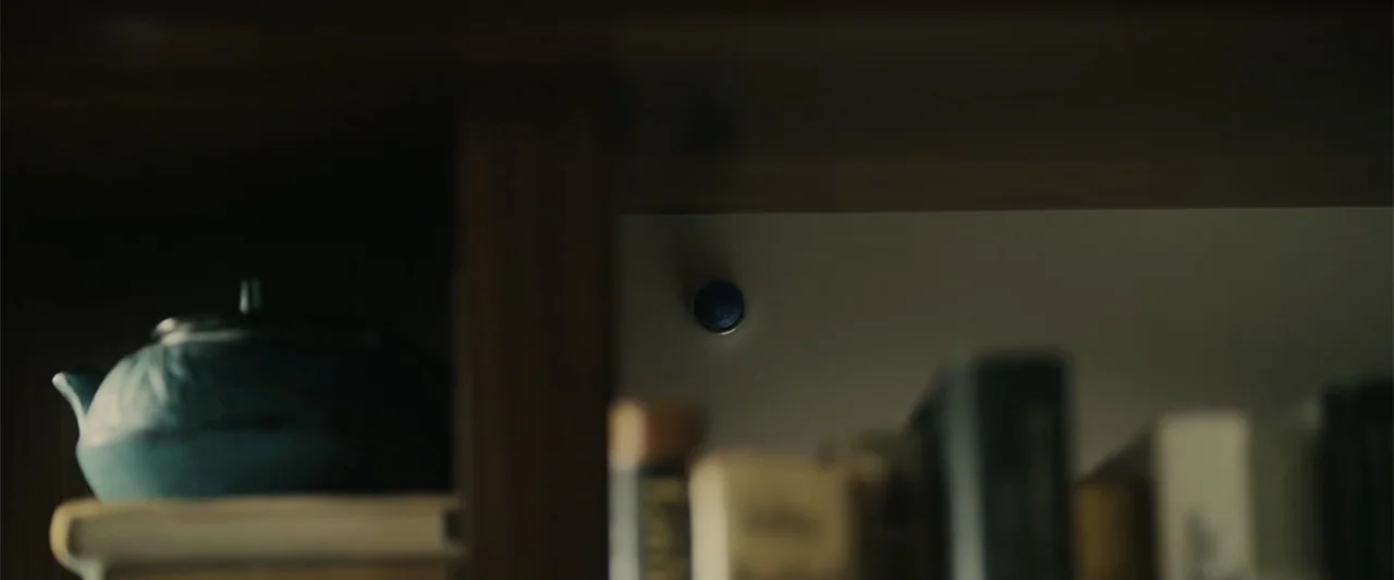 Afraid (2024) movie still: closeup — A dark wooden bookshelf contains a ceramic teapot and several blurred vertical books. A single small blue…