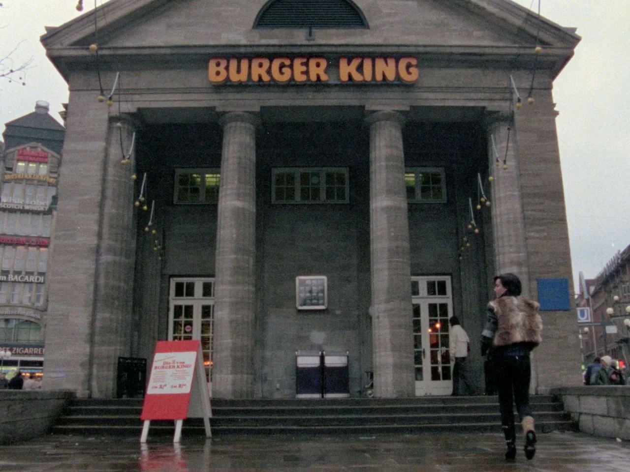 Decoder (1984) movie still: wide — A stone building with large columns houses a Burger King fast food outlet. A woman walks toward the entrance…