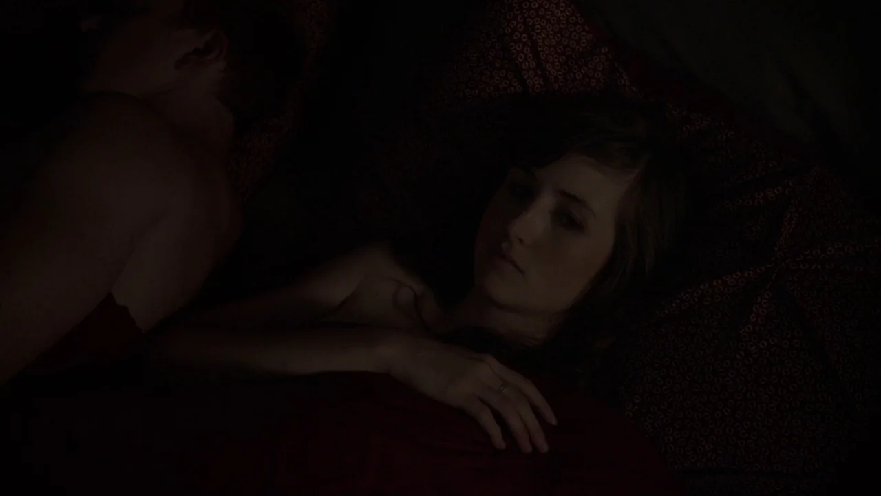 The Heart Machine (2014) movie still: medium — A dark bedroom shows a couple lying together in a bed with patterned sheets. The woman is awake and…