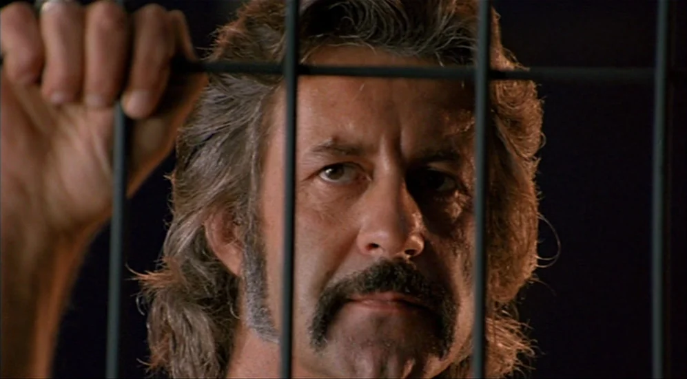 Roselyne and the Lions (1989) movie still: closeup — A man stands behind metal bars in a dark room. He stares forward while gripping one of the…