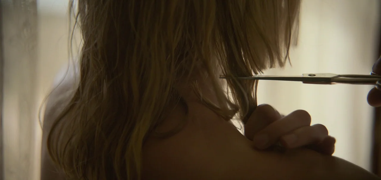 Foe (2023) movie still: closeup — A close view of a person with damp blonde hair viewed from the back. A hand holds silver scissors near the nape of…